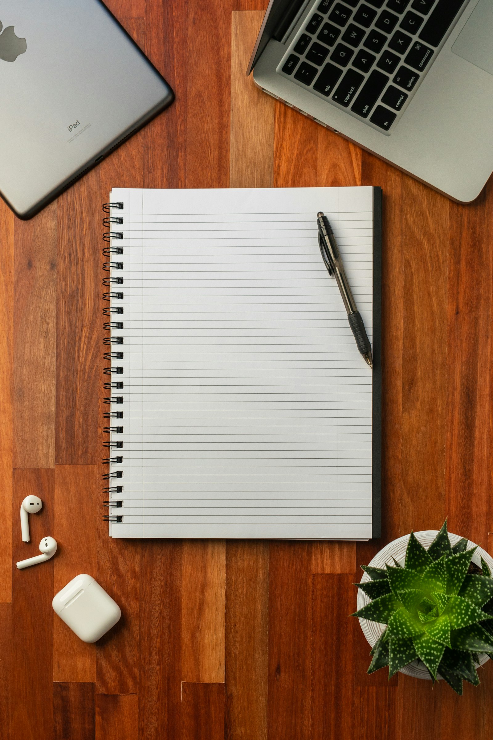 A notebook and pen on a wooden desk between a laptop and tablet