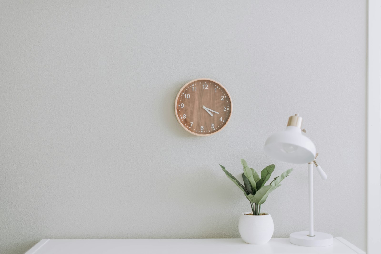 A wall clock next to a desk lamp and small plant on a white desk
