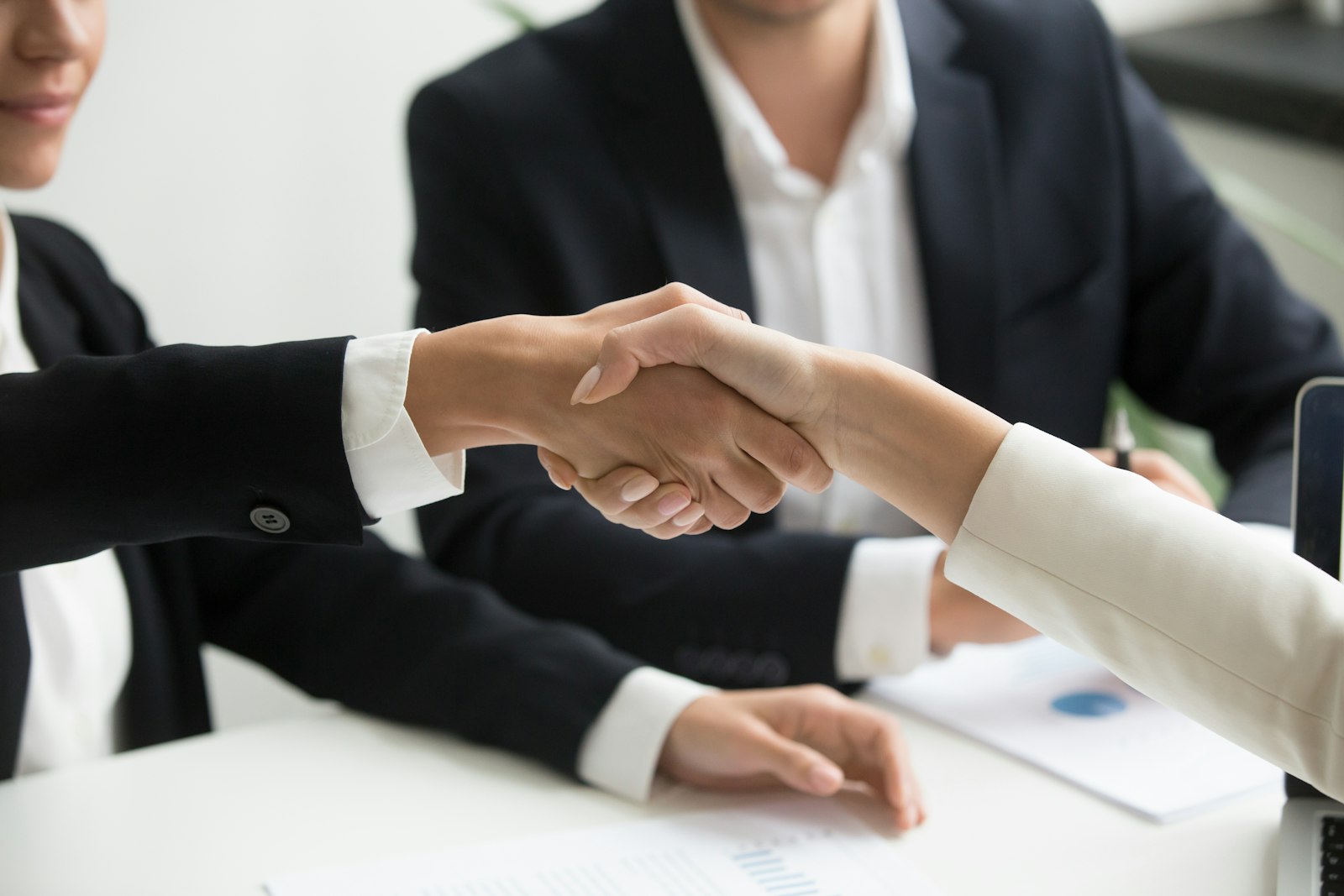Two people shaking hands across a desk in front of a laptop