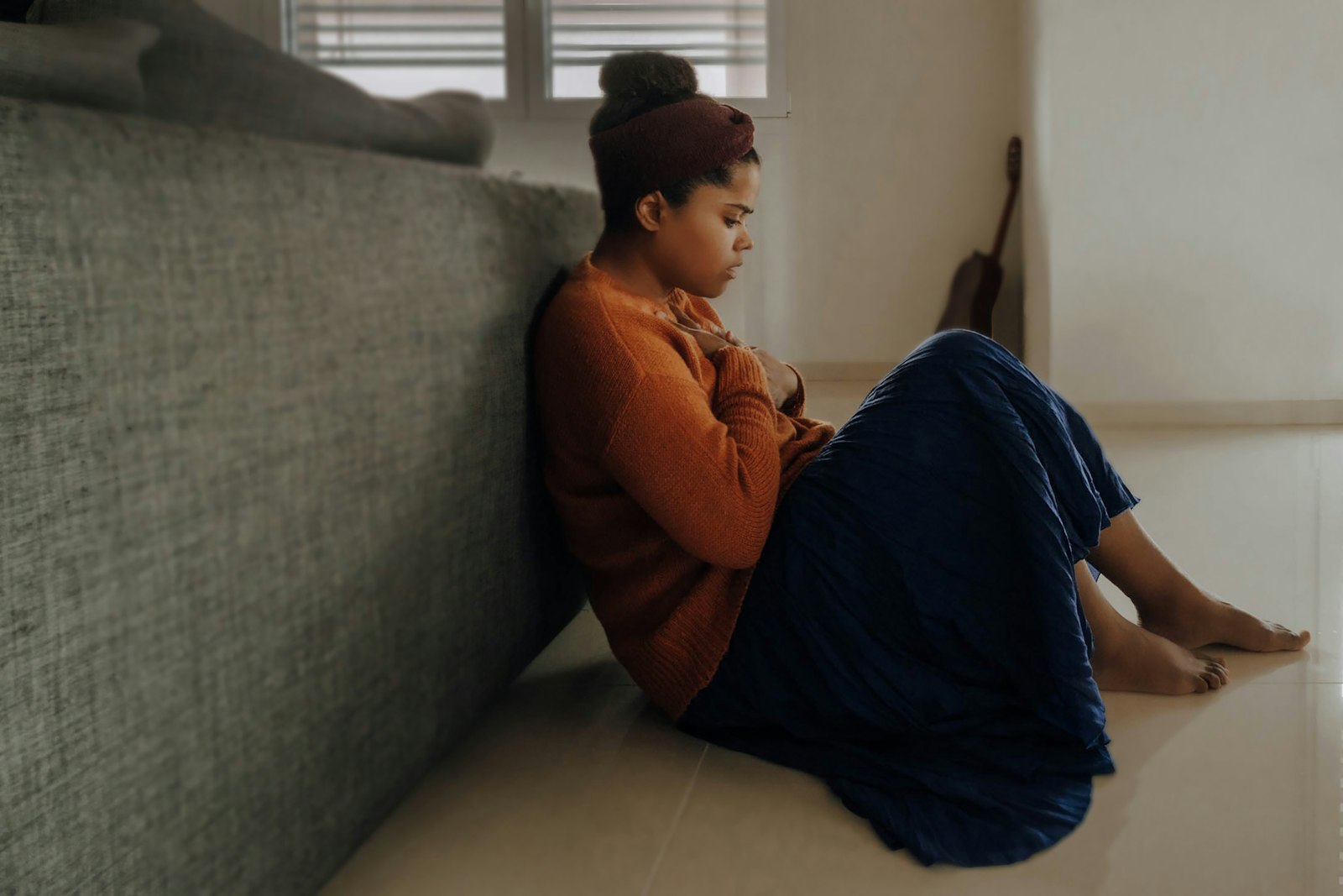 A person sitting on the floor leaning against a couch, looking down
